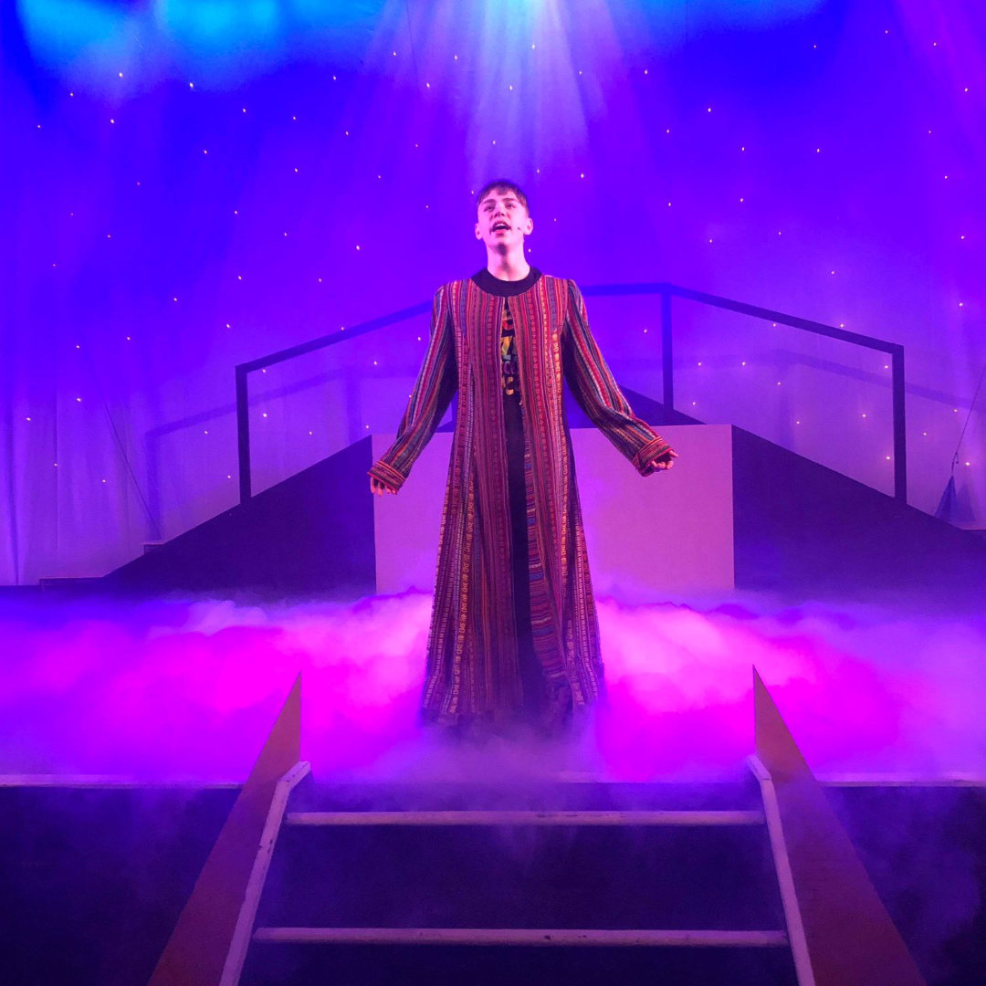 Student dressed in a technicolour coat portraying Joseph on stage