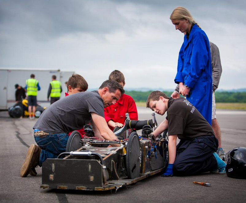Students work on a single seat electric race car by the side of a race track